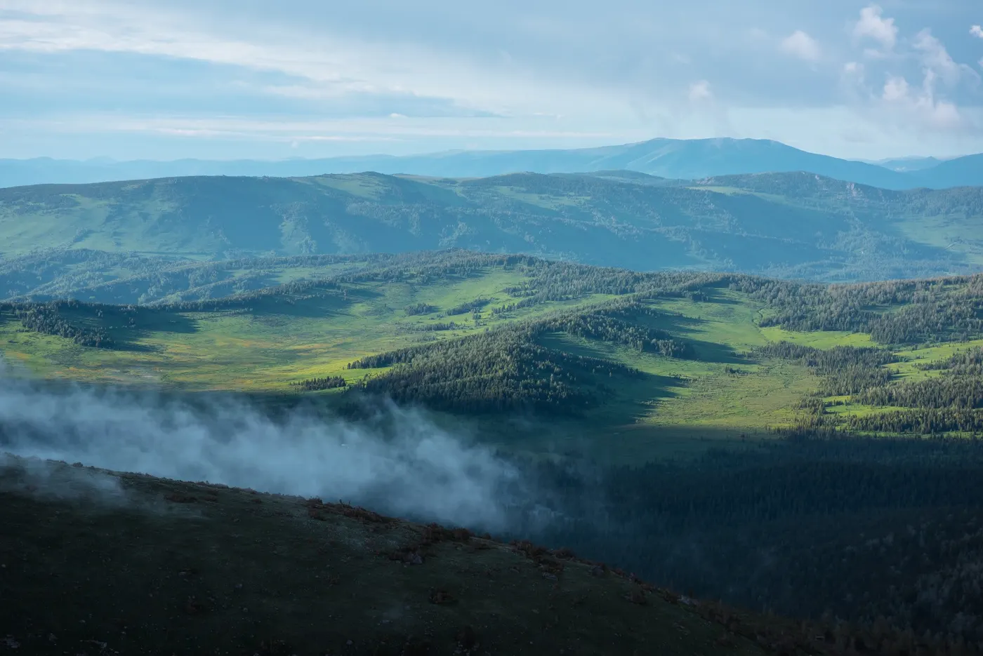 Aerial view of low clouds drifting over green hills and layered mountain silhouettes